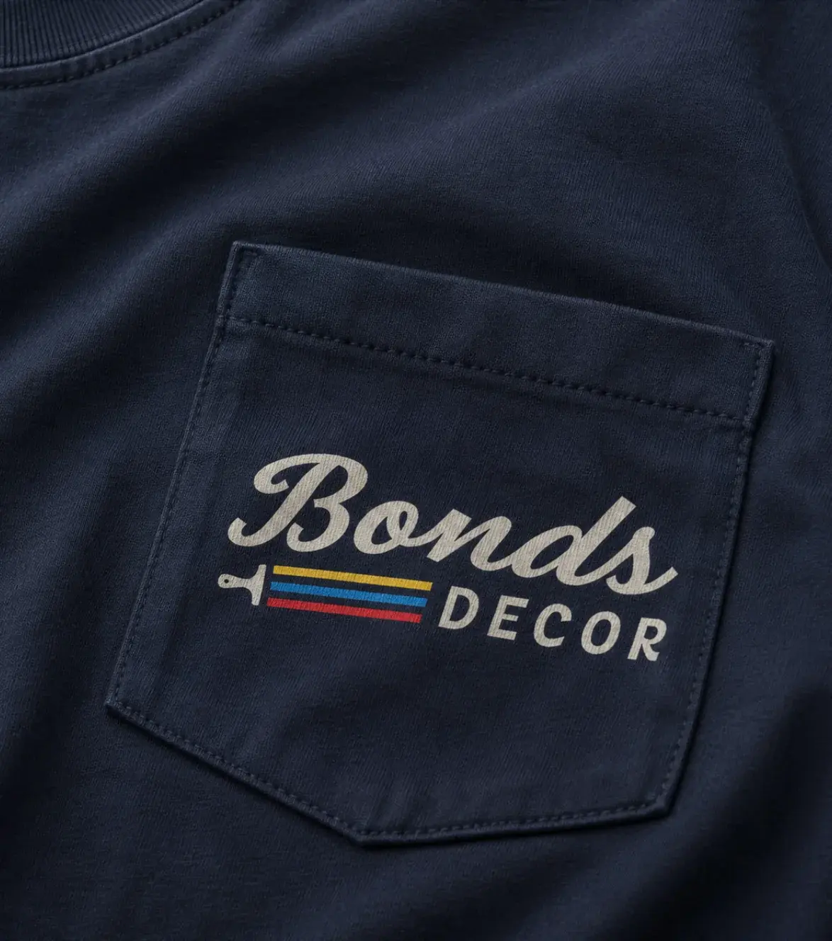 A navy pocket tee, the Bonds Decor script logo printed on the pocket in cream above a paintbrush trailing yellow, blue and red stripes.