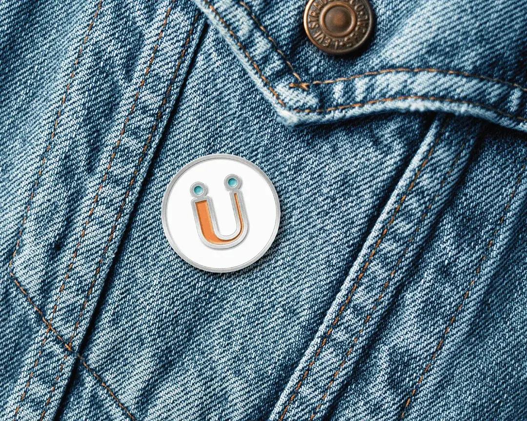 A white enamel pin of the Rückify ü smiley, its orange letter between two teal dots, fastened to the chest pocket of a light denim jacket.