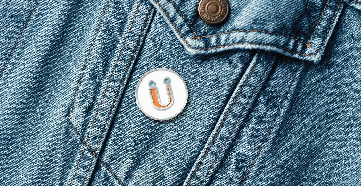 A white enamel pin of the Rückify ü smiley fastened to the pocket of a denim jacket.
