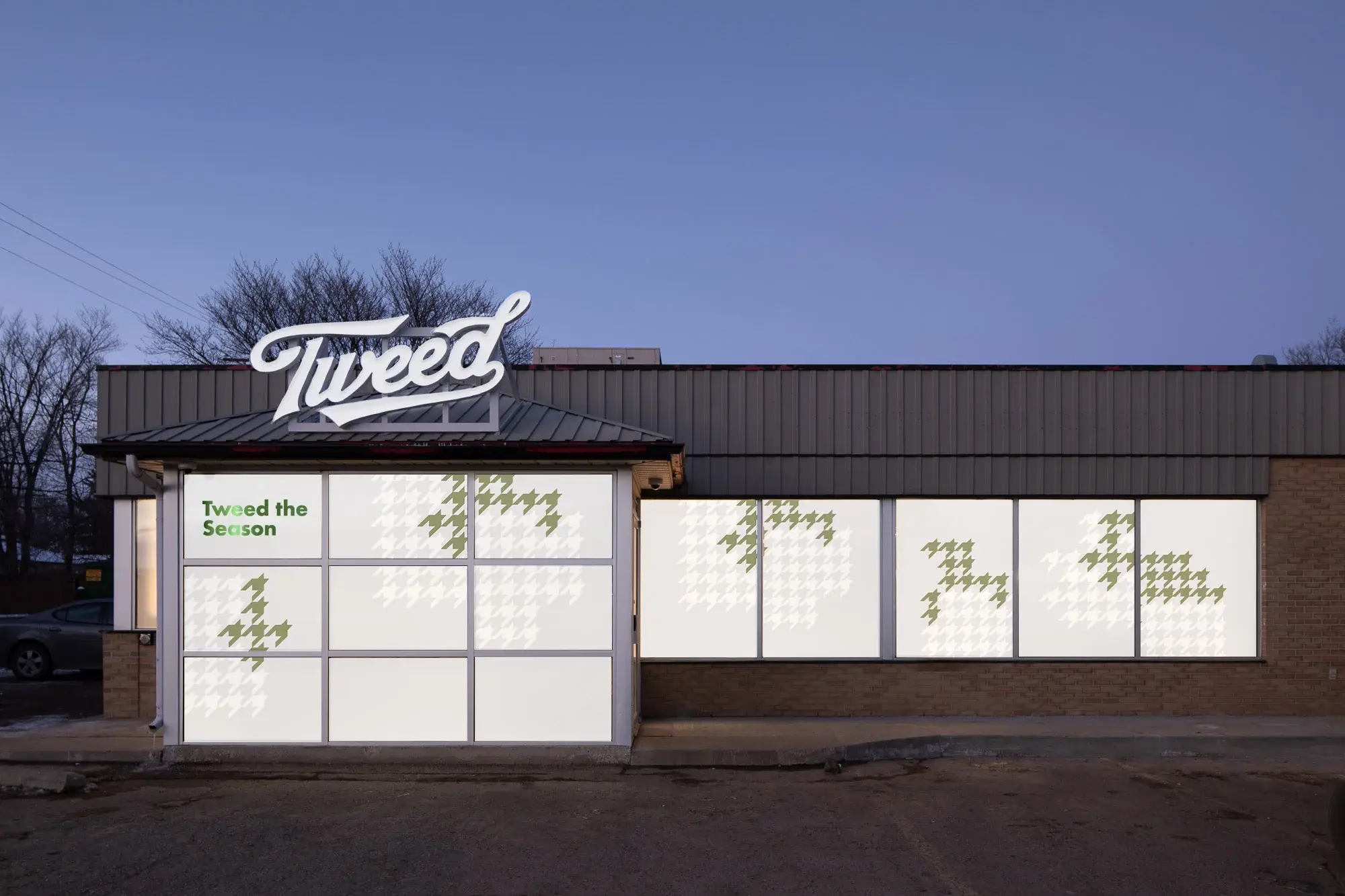 A Tweed storefront at dusk, its windows lit and dressed with the Tweed the Season campaign in pale green.